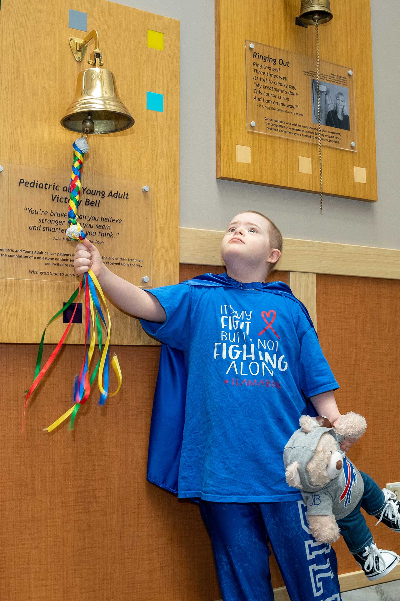 A young child, donning a blue cape and T-shirt, rings a golden victory bell mounted on a wall. Clutching a stuffed bear with determination, the T-shirt boldly declares, "It's a fight, but I'm not fighting alone." This moment embodies the spirit of hope—it's why we ride for change.