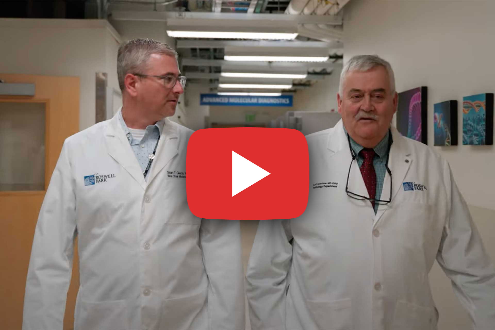 Two men in white lab coats stroll down a hallway, immersed in conversation within a research facility adorned with scientific images and signs. A red play button icon hints at video content titled "Why We Ride," seamlessly blending curiosity with discovery in this intriguing snapshot.