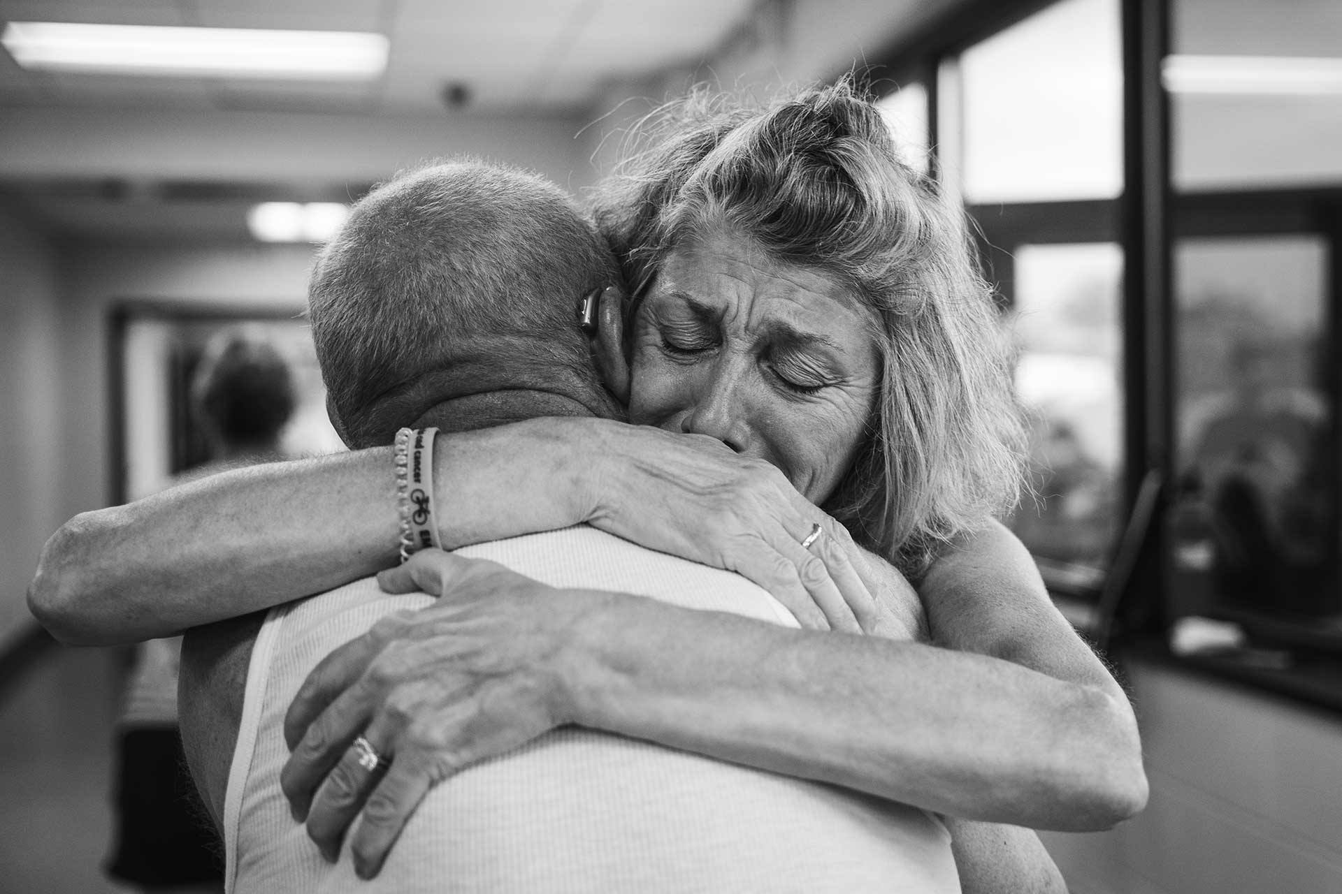 An emotional black and white photo of a woman with tears in her eyes hugging a man. They are indoors, and the woman appears deeply moved, clutching the man tightly.