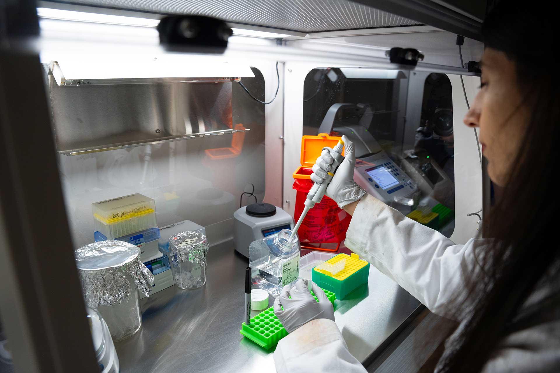 A scientist in a lab coat uses a pipette at a sterile laboratory workstation surrounded by lab equipment and containers. Various colored racks and trays are organized around the workspace, hinting at active scientific research.