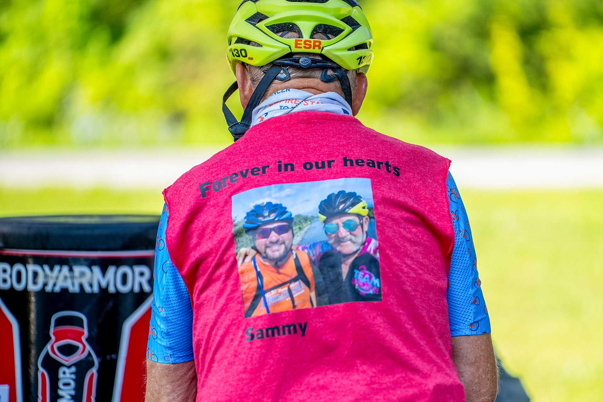 A cyclist wearing a helmet and a red vest with a photo and the words "Forever in our hearts Sammy" printed on the back, standing outdoors near a BODYARMOR drink station.
