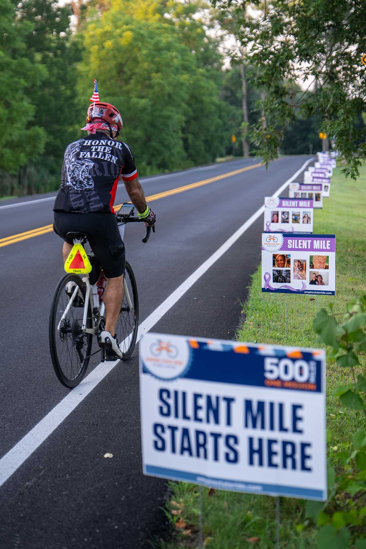 A cyclist rides along a rural road lined with signs featuring photos and text. The nearest sign reads, "SILENT MILE STARTS HERE." The cyclist’s jersey says "HONOR THE FALLEN." The scene is green and peaceful.