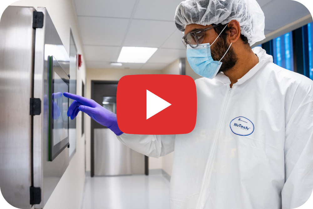 A person in protective lab clothing, gloves, hairnet, and face mask uses a touchscreen in a cleanroom. A large red play button symbol is overlaid in the center of the image.