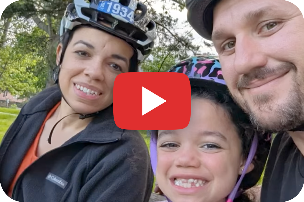 A woman, a young girl, and a man, all wearing bike helmets, smile at the camera outdoors. The image has a large play button over it, suggesting it is a video thumbnail.